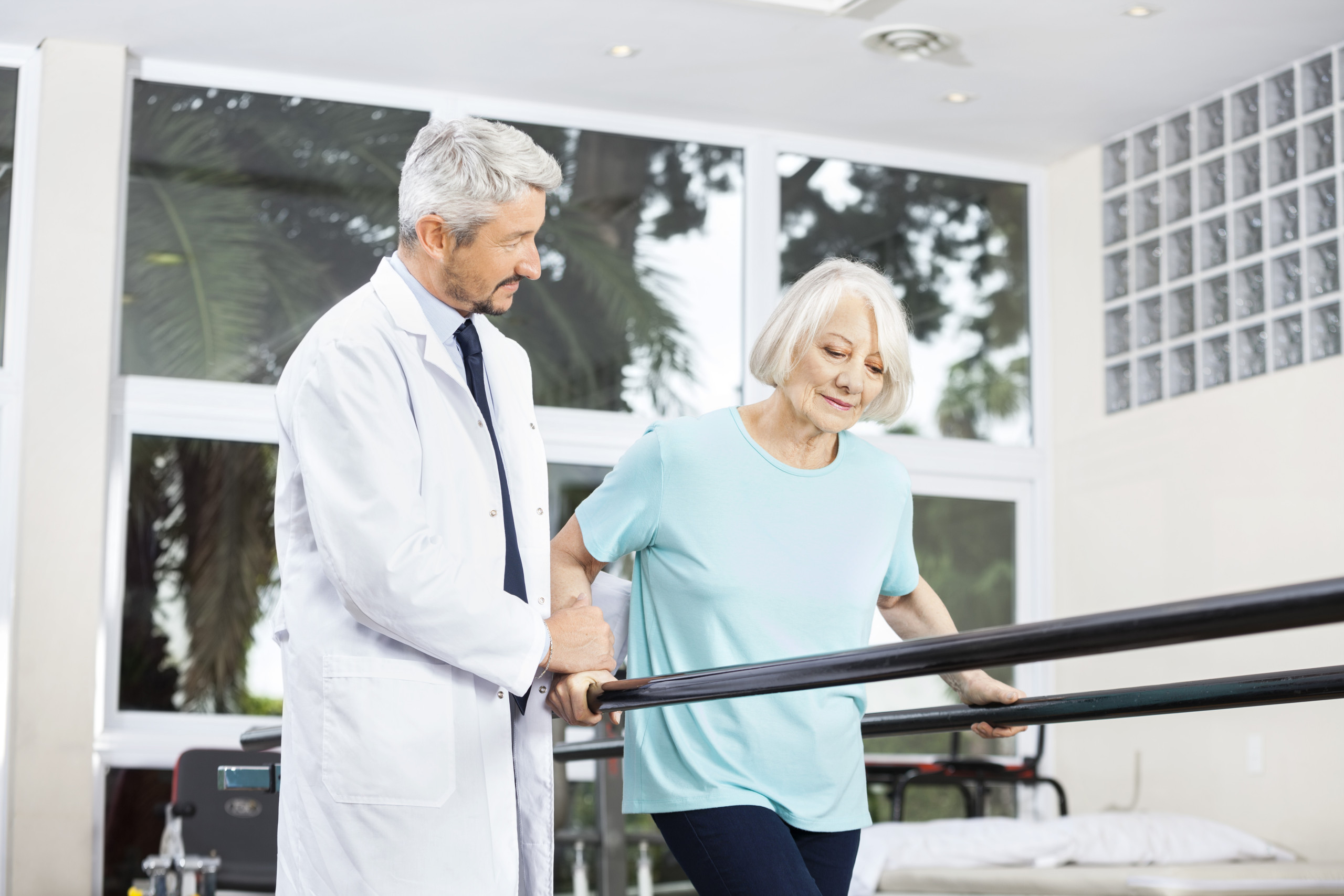 Doctor,Looking,At,Senior,Woman,Walking,Between,Parallel,Bars